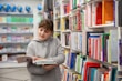 A young person in a gray hoodie is holding and reading a book in a well-organized bookstore or library. Shelves filled with colorful books are visible, creating a vibrant and scholarly atmosphere. In the background, several globes are displayed on a shelf.