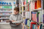 A young person in a gray hoodie is holding and reading a book in a well-organized bookstore or library. Shelves filled with colorful books are visible, creating a vibrant and scholarly atmosphere. In the background, several globes are displayed on a shelf.