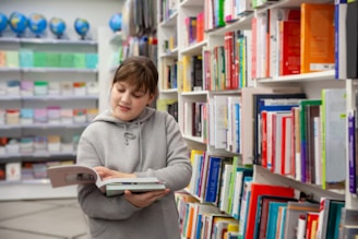 A young person in a gray hoodie is holding and reading a book in a well-organized bookstore or library. Shelves filled with colorful books are visible, creating a vibrant and scholarly atmosphere. In the background, several globes are displayed on a shelf.