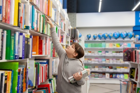 A young person in a gray hoodie is reaching for a book on a tall shelf in a bookstore or library. They are holding multiple books in their other arm, indicating they are actively selecting interesting titles. Shelves filled with colorful books line the background, accompanied by a row of globes and stationary items on display.