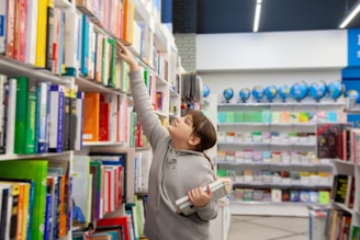 A young person in a gray hoodie is reaching for a book on a tall shelf in a bookstore or library. They are holding multiple books in their other arm, indicating they are actively selecting interesting titles. Shelves filled with colorful books line the background, accompanied by a row of globes and stationary items on display.
