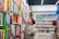 A young person in a gray hoodie is reaching for a book on a tall shelf in a bookstore or library. They are holding multiple books in their other arm, indicating they are actively selecting interesting titles. Shelves filled with colorful books line the background, accompanied by a row of globes and stationary items on display.