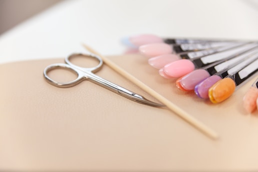 A set of manicure tools including a pair of scissors, a wooden cuticle stick, and a display of various nail polish colors on a beige surface. The nail polishes range from pinks and purples to an orange hue, displayed on sample swatches.