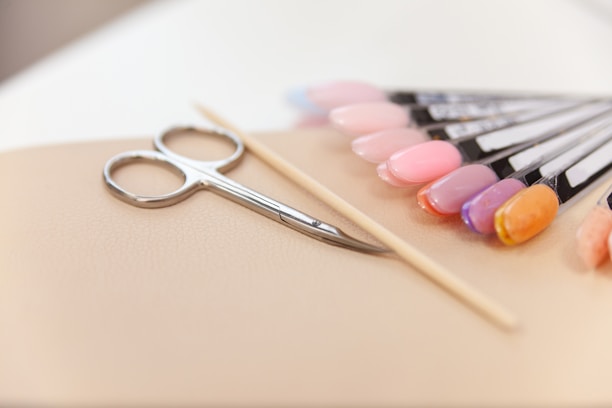 A set of manicure tools including a pair of scissors, a wooden cuticle stick, and a display of various nail polish colors on a beige surface. The nail polishes range from pinks and purples to an orange hue, displayed on sample swatches.