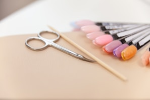 A set of manicure tools including a pair of scissors, a wooden cuticle stick, and a display of various nail polish colors on a beige surface. The nail polishes range from pinks and purples to an orange hue, displayed on sample swatches.