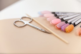 A set of manicure tools including a pair of scissors, a wooden cuticle stick, and a display of various nail polish colors on a beige surface. The nail polishes range from pinks and purples to an orange hue, displayed on sample swatches.