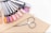 A collection of nail polish color samples displayed on nail swatches fan out over a beige surface. Nearby, a wooden cuticle stick and a small pair of nail scissors are placed. The color samples range from pastels to vibrant hues.