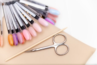 A collection of nail polish color samples displayed on nail swatches fan out over a beige surface. Nearby, a wooden cuticle stick and a small pair of nail scissors are placed. The color samples range from pastels to vibrant hues.