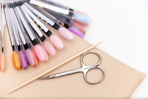 A collection of nail polish color samples displayed on nail swatches fan out over a beige surface. Nearby, a wooden cuticle stick and a small pair of nail scissors are placed. The color samples range from pastels to vibrant hues.