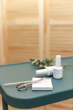 A teal-colored table holds a pair of metal scissors, a wooden cuticle stick, and two bottles of nail polish. There is also a small white pad and some greenery in the background, set against a blurred, light brown backdrop.