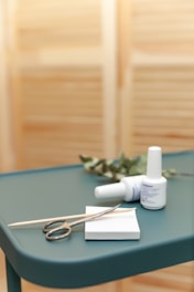 A teal-colored table holds a pair of metal scissors, a wooden cuticle stick, and two bottles of nail polish. There is also a small white pad and some greenery in the background, set against a blurred, light brown backdrop.