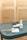 A teal-colored table holds a pair of metal scissors, a wooden cuticle stick, and two bottles of nail polish. There is also a small white pad and some greenery in the background, set against a blurred, light brown backdrop.