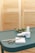 A teal-colored table holds a pair of metal scissors, a wooden cuticle stick, and two bottles of nail polish. There is also a small white pad and some greenery in the background, set against a blurred, light brown backdrop.