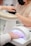 A person is receiving a manicure, with the focus on their hand under a UV nail lamp. Another person wearing a mask is performing the manicure. A beige armrest is visible next to the lamp.