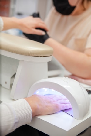 A person is receiving a manicure, with the focus on their hand under a UV nail lamp. Another person wearing a mask is performing the manicure. A beige armrest is visible next to the lamp.