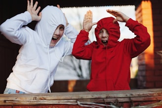 Two children are playfully posing with their hoodie drawstrings pulled tight around their faces, laughing with their hands raised in a playful manner. The child on the left wears a white hoodie, and the one on the right wears a red hoodie. They are standing in a warm outdoor setting, perhaps a backyard or playground, with a background that includes a brick wall.