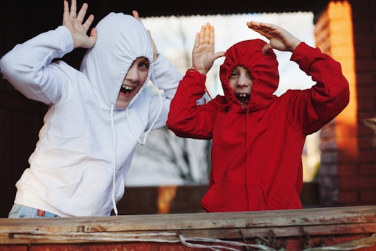 Two children are playfully posing with their hoodie drawstrings pulled tight around their faces, laughing with their hands raised in a playful manner. The child on the left wears a white hoodie, and the one on the right wears a red hoodie. They are standing in a warm outdoor setting, perhaps a backyard or playground, with a background that includes a brick wall.