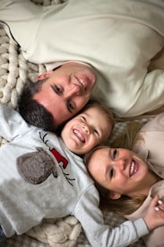 A family of three lies on a cozy, textured blanket, smiling and embracing each other affectionately. The parent and child are positioned head to head in a close-knit arrangement. The child wears a sweater with a playful reindeer design.