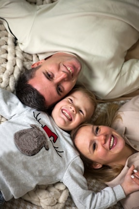 A family of three lies on a cozy, textured blanket, smiling and embracing each other affectionately. The parent and child are positioned head to head in a close-knit arrangement. The child wears a sweater with a playful reindeer design.