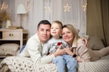 A family of three is lying on a cozy bed, closely embracing each other with smiles on their faces. The room is warmly lit with decorative lights hanging in the background. A bedside table with a lamp, a stack of books, and some decorative items can be seen.