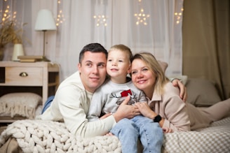A family of three is lying on a cozy bed, closely embracing each other with smiles on their faces. The room is warmly lit with decorative lights hanging in the background. A bedside table with a lamp, a stack of books, and some decorative items can be seen.