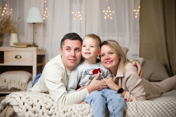 A family of three is lying on a cozy bed, closely embracing each other with smiles on their faces. The room is warmly lit with decorative lights hanging in the background. A bedside table with a lamp, a stack of books, and some decorative items can be seen.