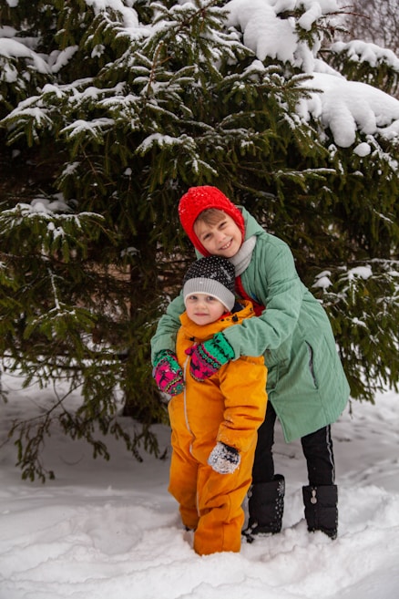 Two children dressed in winter clothing are playing outside in the snow in front of a snow-covered evergreen tree. One child is wearing an orange snowsuit and a grey hat, while the other is in a green coat with a red hat, smiling and embracing the other child. Both have colorful gloves, and the ground is covered with snow.