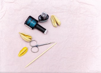 A flat lay featuring nail care items including a black nail polish bottle and a topcoat, two dried seed pods, a metal cuticle pusher, and a stick. The background is a textured light pink surface.