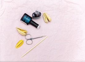 A flat lay featuring nail care items including a black nail polish bottle and a topcoat, two dried seed pods, a metal cuticle pusher, and a stick. The background is a textured light pink surface.