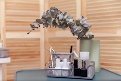 A table surface holds a metal mesh organizer containing various manicure tools, nail polishes, and cosmetic bottles. Behind it, a green vase with eucalyptus branches is placed against a wooden slatted background.