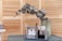 A table surface holds a metal mesh organizer containing various manicure tools, nail polishes, and cosmetic bottles. Behind it, a green vase with eucalyptus branches is placed against a wooden slatted background.