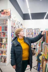 A person in a bookstore reaching for a book on a shelf. Shelves are lined with colorful books, and the store is well-lit with overhead lights. The person is wearing a dark jacket and a yellow top, looking intently at the selection.