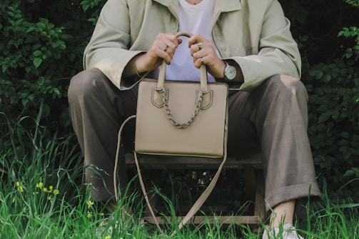 A person is sitting on a wooden stool outdoors, surrounded by green foliage and grass. They are holding a beige handbag with both hands, displaying a casual and relaxed posture. The individual is wearing a light-colored jacket, a white shirt, and brown pants. A watch is visible on their wrist, and the environment gives off a natural, tranquil vibe.