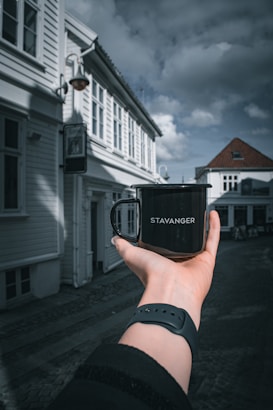 A person holds a black mug with the word 'STAVANGER' printed on it, set against an old European-style street with white wooden buildings and a cloudy sky.