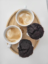 Two cups of cappuccino with frothy milk are placed on a circular wooden tray alongside two chocolate muffins. The arrangement suggests a cozy and inviting atmosphere, perfect for a coffee break.