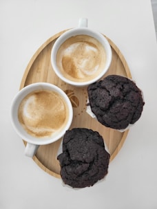 Two cups of cappuccino with frothy milk are placed on a circular wooden tray alongside two chocolate muffins. The arrangement suggests a cozy and inviting atmosphere, perfect for a coffee break.