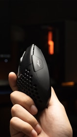 A hand is holding a black ergonomic mouse with perforations on the sides and a logo on the top. The background is dimly lit with an orange light source visible in the distance.