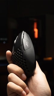 A hand is holding a black ergonomic mouse with perforations on the sides and a logo on the top. The background is dimly lit with an orange light source visible in the distance.