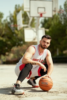 A person wearing a white and red basketball jersey crouches on an outdoor basketball court, holding an orange basketball. Behind them, several basketball hoops are visible against a backdrop of green trees and a clear sky.