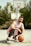A person wearing a white and red basketball jersey crouches on an outdoor basketball court, holding an orange basketball. Behind them, several basketball hoops are visible against a backdrop of green trees and a clear sky.