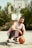 A person wearing a white and red basketball jersey crouches on an outdoor basketball court, holding an orange basketball. Behind them, several basketball hoops are visible against a backdrop of green trees and a clear sky.