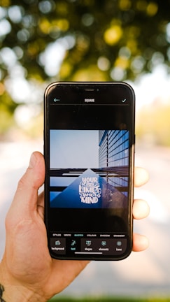 A hand is holding a smartphone displaying an image editing app. The app shows a photo of a building with reflective glass panels and the words 'Your only limit is your mind' overlaid on it. The background is out of focus, featuring greenery and dappled sunlight.