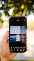 A hand is holding a smartphone displaying an image editing app. The app shows a photo of a building with reflective glass panels and the words 'Your only limit is your mind' overlaid on it. The background is out of focus, featuring greenery and dappled sunlight.