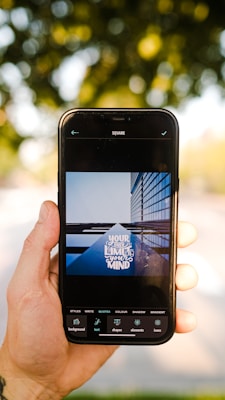 A hand is holding a smartphone displaying an image editing app. The app shows a photo of a building with reflective glass panels and the words 'Your only limit is your mind' overlaid on it. The background is out of focus, featuring greenery and dappled sunlight.