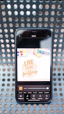 A smartphone displaying a digital design application with a bright screen showing the text 'Live Your Passion' in orange on a white background. The phone rests on a surface with a perforated pattern, and various design tools are visible at the bottom of the screen.