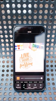 A smartphone displaying a digital design application with a bright screen showing the text 'Live Your Passion' in orange on a white background. The phone rests on a surface with a perforated pattern, and various design tools are visible at the bottom of the screen.