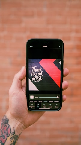 A hand is holding a smartphone displaying an image editing app. The screen shows a design with the text 'Think like an artist' in white on a dark background with red and black geometric shapes. The background features a blurred, rustic brick wall.