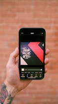 A hand is holding a smartphone displaying an image editing app. The screen shows a design with the text 'Think like an artist' in white on a dark background with red and black geometric shapes. The background features a blurred, rustic brick wall.