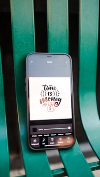 A smartphone displaying a digital art design with the phrase 'time is money' along with icons for currency and a timepiece. The phone is placed on a green metal chair or bench.