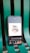 A smartphone displaying a digital art design with the phrase 'time is money' along with icons for currency and a timepiece. The phone is placed on a green metal chair or bench.